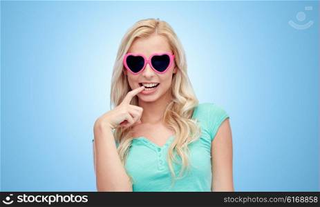 emotions, expressions, summer and people concept - smiling young woman or teenage girl in heart shape sunglasses over blue background