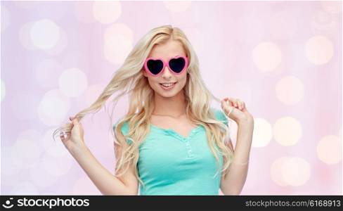 emotions, expressions, summer and people concept - smiling young woman or teenage girl in heart shape sunglasses holding her strand of hair over pink holidays lights background