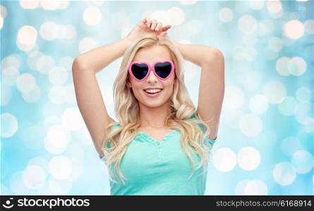 emotions, expressions, summer and people concept - smiling young woman or teenage girl in heart shape sunglasses over blue holidays lights background
