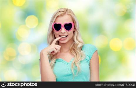 emotions, expressions, summer and people concept - smiling young woman or teenage girl in heart shape sunglasses over summer green lights background