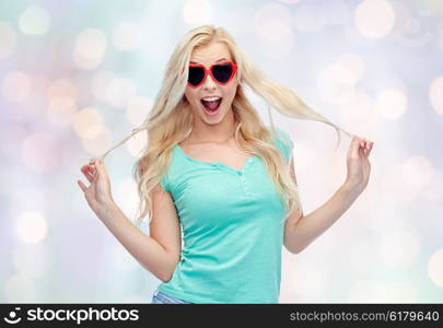 emotions, expressions, summer and people concept - smiling young woman or teenage girl in heart shape sunglasses holding her strand of hair over holidays lights background