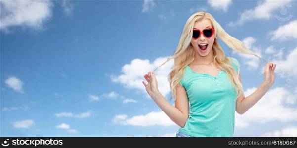 emotions, expressions, summer and people concept - smiling young woman or teenage girl in heart shape sunglasses holding her strand of hair over blue sky and clouds background