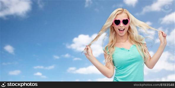 emotions, expressions, summer and people concept - smiling young woman or teenage girl in sunglasses holding her strand of hair over blue sky and clouds background