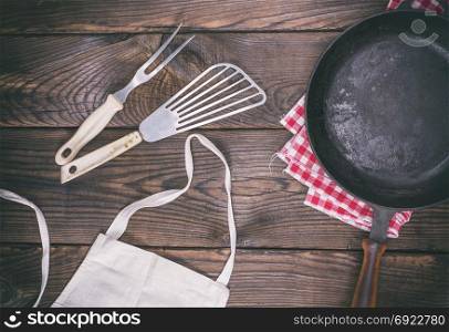 empty black cast-iron frying pan and cooking utensils on a brown wooden background, top view, vintage toning