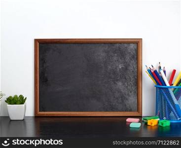empty black chalk board, multi-colored pencils in a blue metal stand on a black table, concept back to school