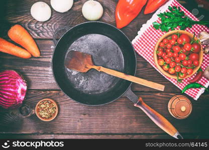 Empty black frying pan with fresh vegetables on a wooden table, top view