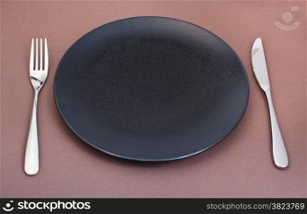 empty black plate with fork and knife set on brown background