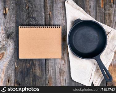 empty black round frying pan with handle and paper notebook on a wooden table, top view