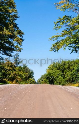 Empty country dirt road hill rising against empty blue sky and surrounding trees.