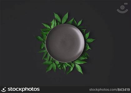 Empty grey plate on black background, surrounded by green leaves. Flat lay of empty dish. Food mockup concept. Empty tableware and copy space.