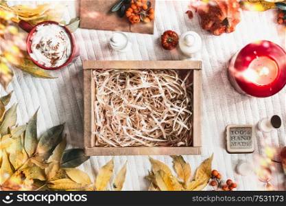 Empty kraft paper box on white blanket with autumn branches and fall leaves, cup of cappuccino, cosmetic products, burning candle and rowan. Zero waste, eco friendly packaging. Beauty box. Top view