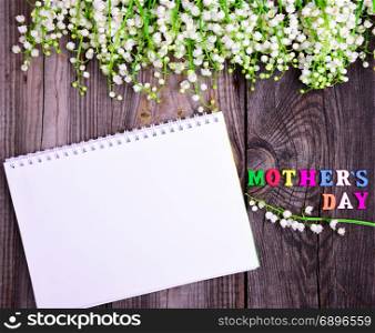empty open white notebook and a congratulatory inscription with a mother&rsquo;s day on a gray wooden background, at the top a bouquet of blossoming lilies of the valley