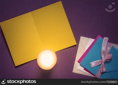Empty paper message card, a lit candle on it and a pile of multicolored envelopes tied with ribbon and bow, on a purple wooden background.