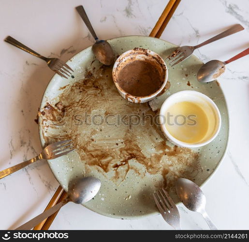 Empty plate with food leftovers from dessert meal, spoons and forks on marble table. Dirty plate with chocolate sauce, chocolate and vanilla ice cream leftovers in ramekin bowl.