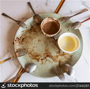 Empty plate with food leftovers from dessert meal, spoons and forks on marble table. Dirty plate with chocolate sauce, chocolate and vanilla ice cream leftovers in ramekin bowl.