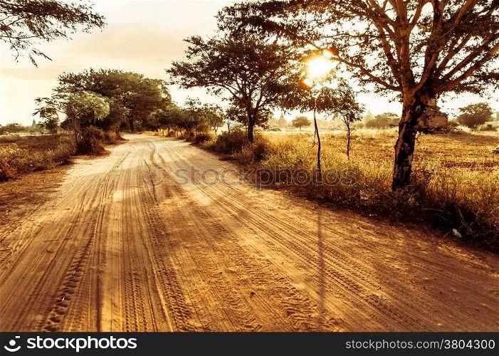 Empty road going through rural landscape under sunset sky with sun beams. Dry season in southeast asia, Myanmar (Burma). Nature background in vintage style