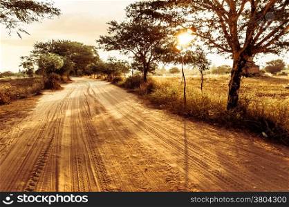 Empty road going through rural landscape under sunset sky with sun beams. Dry season in southeast asia, Myanmar (Burma). Nature background in vintage style