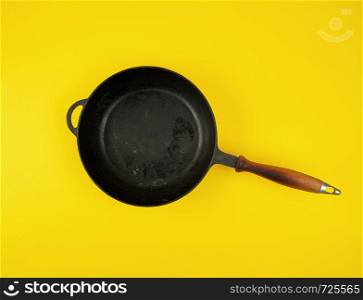 empty round black cast-iron frying pan with wooden handle on yellow background, flat lay