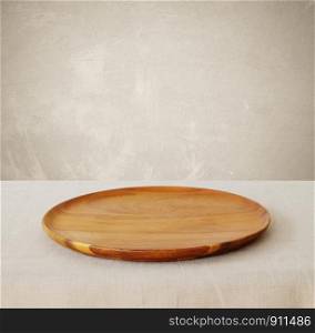 Empty round wooden tray on linen tablecloth over brown cement wall background, kitchen utensil, food display montage