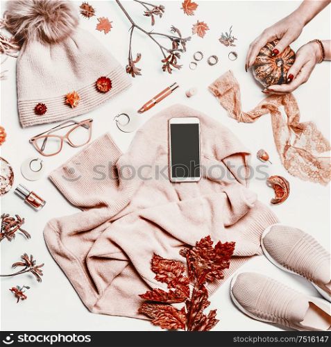 Empty screen smartphone on autumn feminine clothes: knitted sweater, hat and sneakers shoes on white desktop background with female hands , pumpkins, make up cosmetics, glasses and fall leaves