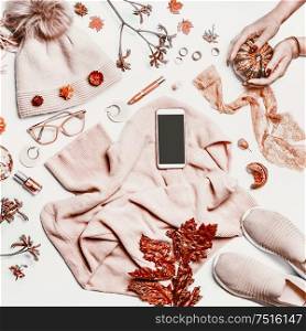 Empty screen smartphone on autumn feminine clothes: knitted sweater, hat and sneakers shoes on white desktop background with female hands , pumpkins, make up cosmetics, glasses and fall leaves