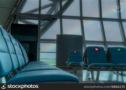 Empty seat in departure lounge at airport terminal. Distance for one seat keep distance to protect coronavirus and passenger social distancing for safety. Seen rainbow on the sky through glass window.