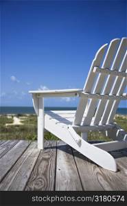 Empty white adirondack chair on wooden deck facing beach.