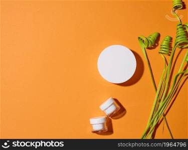 empty white glass jars with plastic lid on orange background with green decor. Packaging for cream, gel, serum, advertising and product promotion. Mock up