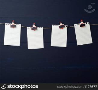 empty white notebook paper sheets hanging on decorative holiday clothespins, blue wooden background. Clean sheet, template. Design element.
