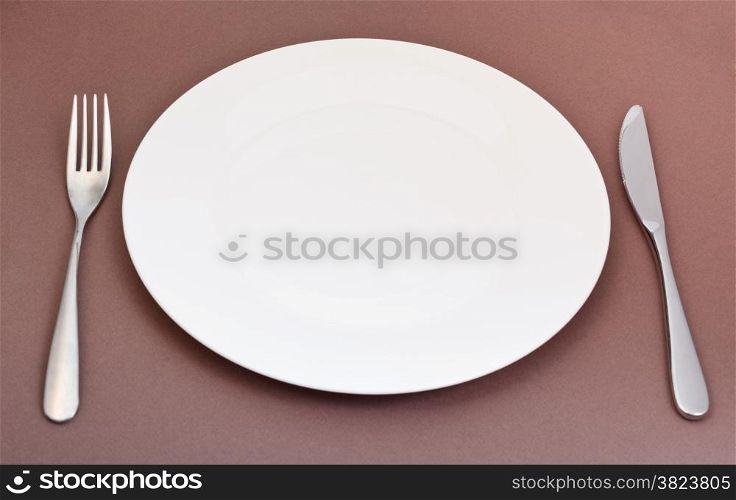 empty white porcelain plate with fork and knife set on brown background