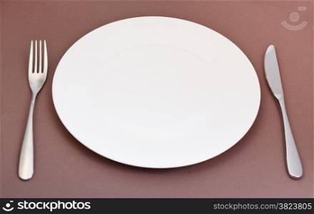 empty white porcelain plate with fork and knife set on brown background
