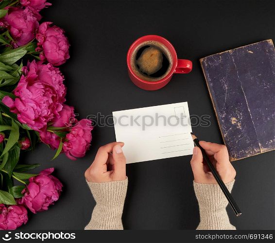 empty white postcard and two female hands, on the table a bouquet of red peonies and a ceramic cup of coffee, top view