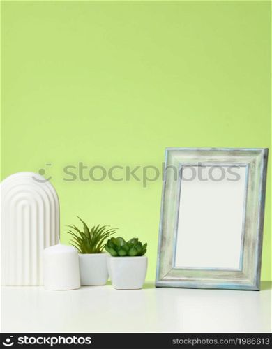 empty white wooden photo frame and flowerpots with plants on white table, green background