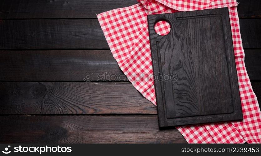 empty wooden cutting board and folded red and white cotton kitchen napkin on a wooden brown background, top view, copy space