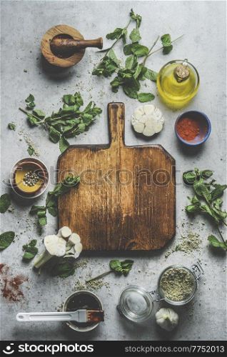 Empty wooden cutting board on grey concrete kitchen table with herbs ,spices, garlic,oil and mortar and pestle. Preparing homemade marinade for meat or vegetables at home.Top view . Frame.