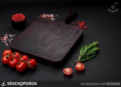 Empty wooden cutting board with cherry tomatoes, spices, salt and herbs on dark concrete background