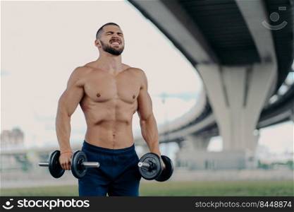 Endurance and power concept. Motivated bearded European man raises barbells puts all efforts in lifting heavy weight clenches teeth poses with sport equipment under bridge, has muscular body