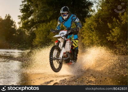 Enduro rider crossing water and muddy terrain against a beautiful sunset.
