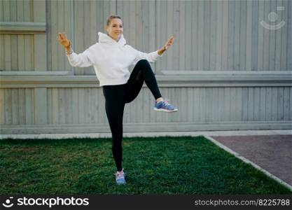 Energetic sportwoman has workout routine poses on one legs demonstrates her stamina warms up before jogging dressed in hoodie leggings and trainers trains outdoors. Sport and motivation concept