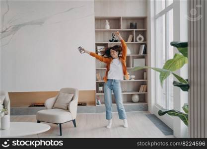 Energetic young woman dancing in living room alone by music from smartphone. Joyful female homeowner dances holding phone, having fun, enjoying musical sound in modern apartment at home interior.. Energetic woman dancing in living room by music from smartphone in modern apartment home interior