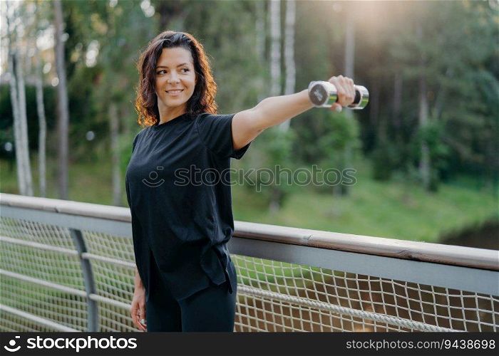 Energized sportswoman stretches arms with dumbbell on a bridge at sunrise. Active wear, focused on biceps training, embraces the morning workout.