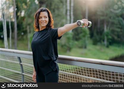 Energized sportswoman stretches arms with dumbbell on a bridge at sunrise. Active wear, focused on biceps training, embraces the morning workout.
