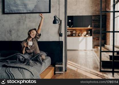 Energy drink. Young beautiful positive european woman sitting on bed stretching with her hand up while holding cup of coffee with another hand, trying to wake up before going to shower in morning. Young beautiful woman sitting on bed stretching with her hand up while holding cup of coffee