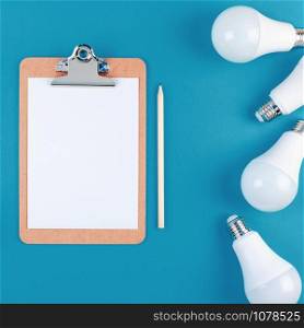 Energy-saving and eco-friendly life in conceptual composition. Creative top view flat lay of LED light bulbs frame and blank paper in clipboard with copy space on blue background minimal style