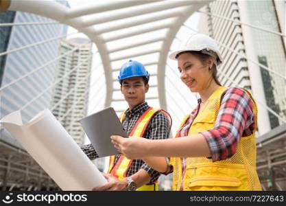 Engineer Asian man and American woman check project plan by company application from digital tablet and paper blueprint. Hapy teamwork at construction site. Win bid auction from internet.