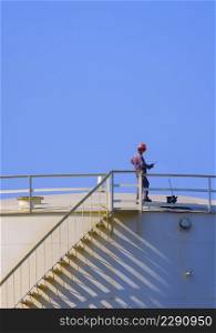 Engineer checking oil quality on top of oil fuel storage tank against blue clear sky in vertical frame