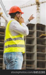 Engineer in red hardhat talking by phone and pointing at building site