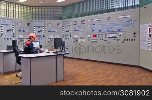 engineer looks at monitor and writes down work log near main control panel of gas compressor station, wide angle