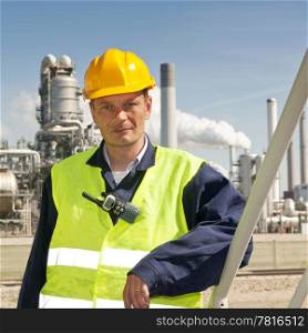 Engineer poses casually in front of a refinery, wearing a safety vest and hard hat