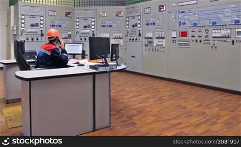 engineer sits and talks on phone near main control panel of gas compressor station, then looks at monitor while making notes in registry